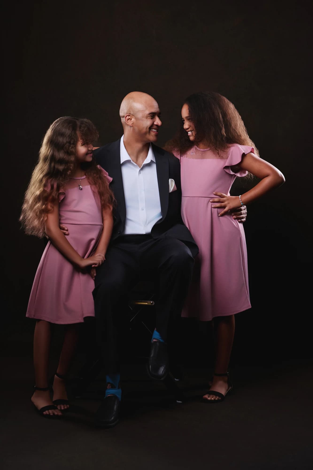 A father seated with his two daughters in matching rose-colored dresses