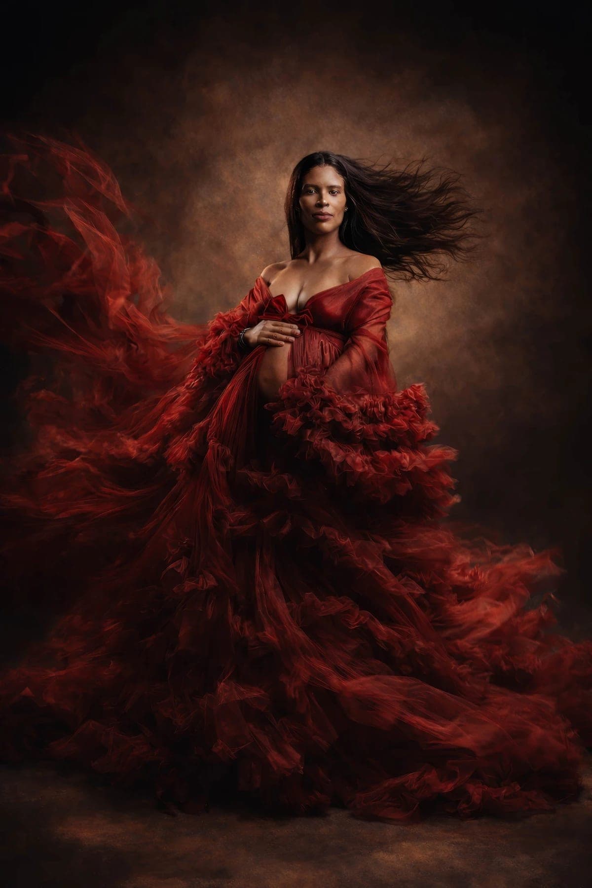 Pregnant woman in flowing red fabric gown holds her belly, hair blowing against dark background.