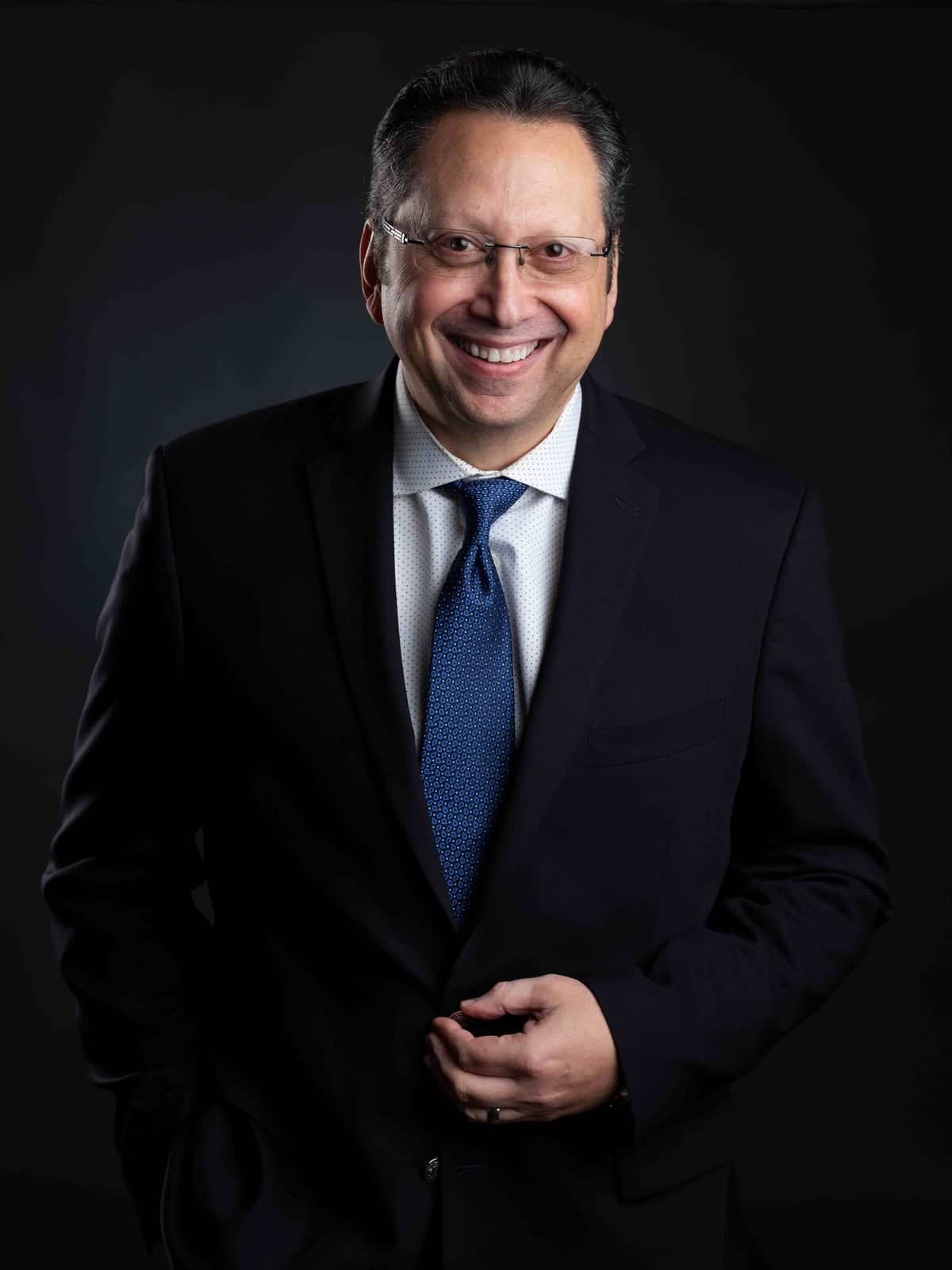 A smiling man wearing glasses and a navy suit in the studio