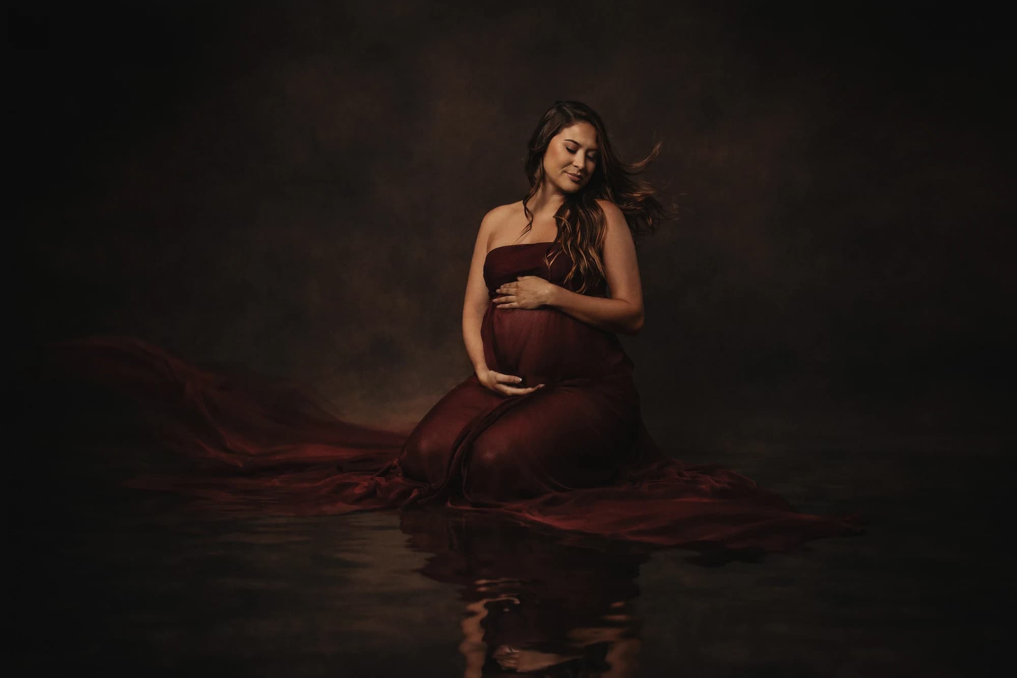 An expectant mother in a burgundy gown seated above a softly lit reflection