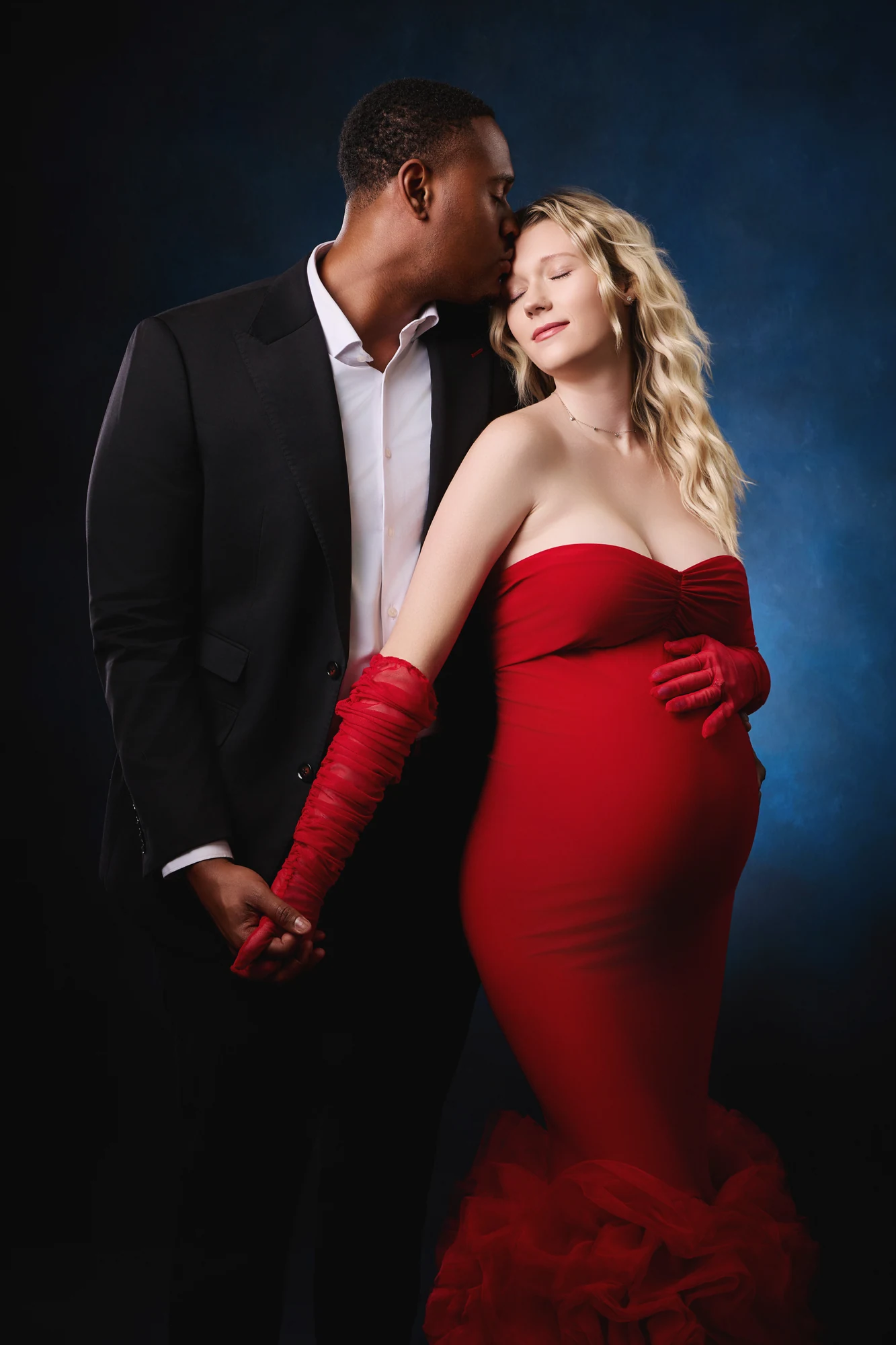 Expectant parents holding hands, with a red gown against a blue studio backdrop